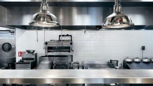 Clean commercial kitchen with stainless steel appliances, overhead hood system, industrial lighting, and organized cooking equipment, demonstrating proper maintenance and degreasing.